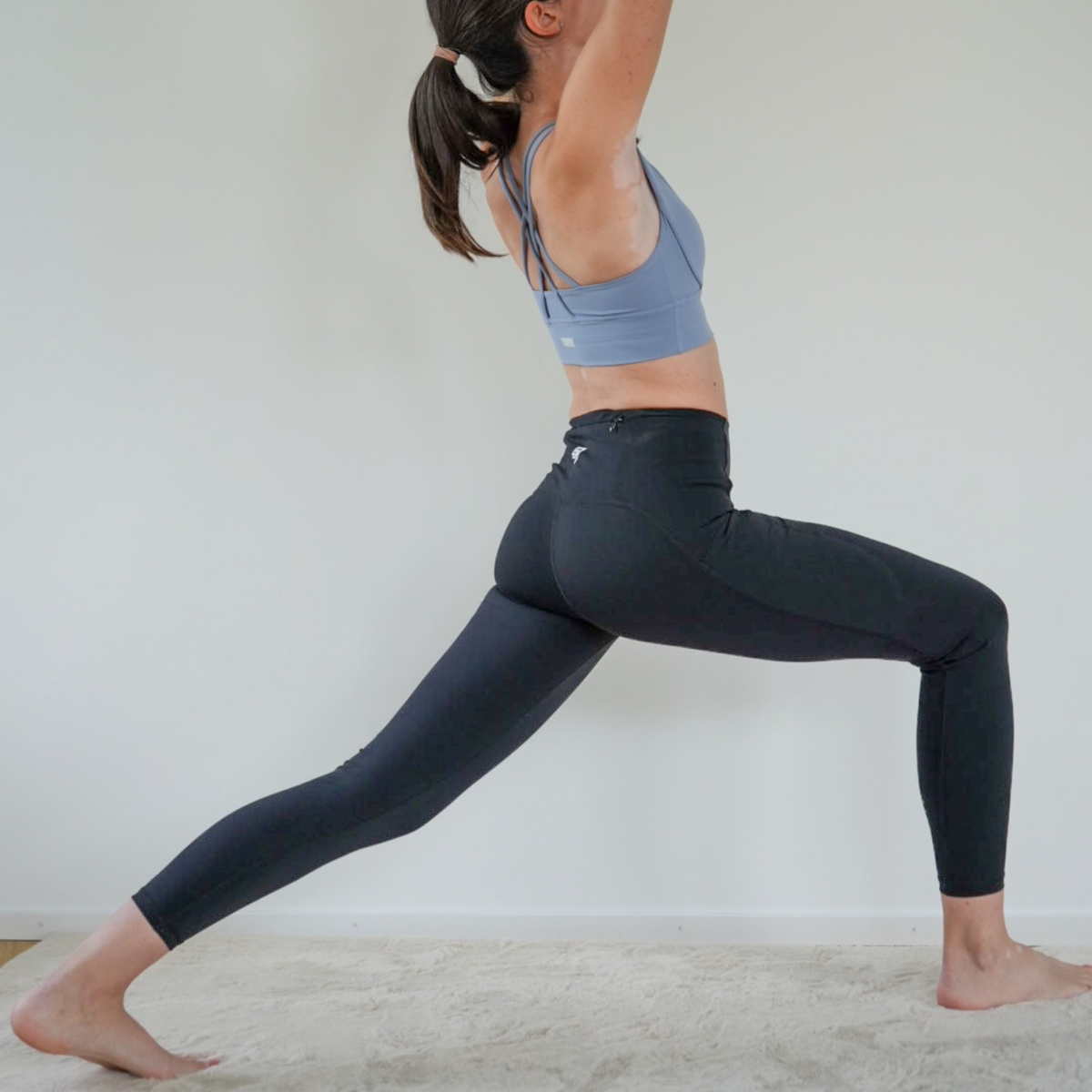 A woman in a yoga pose wearing a light blue sports bra and black high-waisted leggings, standing on a light-colored rug against a plain white wall.