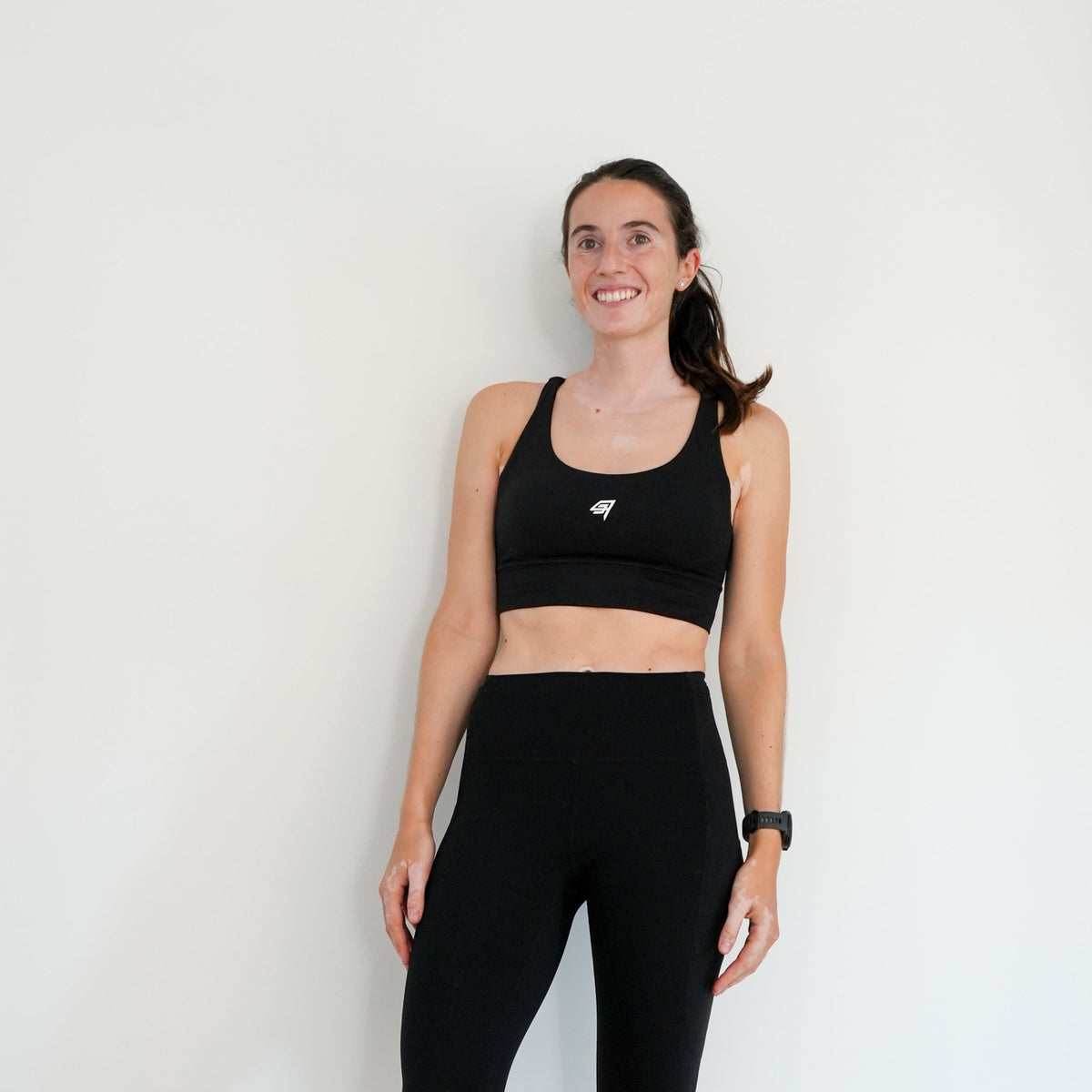 A woman modeling a matching black activewear set consisting of a sports bra and high-waisted leggings, standing against a plain white wall.