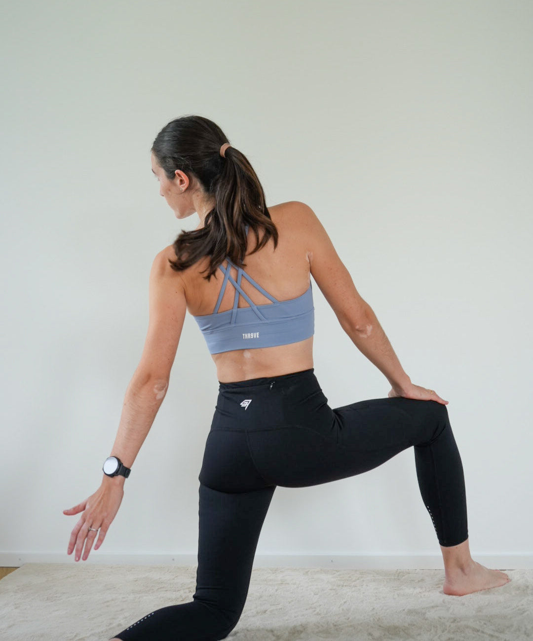 A woman in a yoga pose wearing a light blue sports bra and black leggings, photographed from behind in a minimalist room with a light gray wall and beige floor.