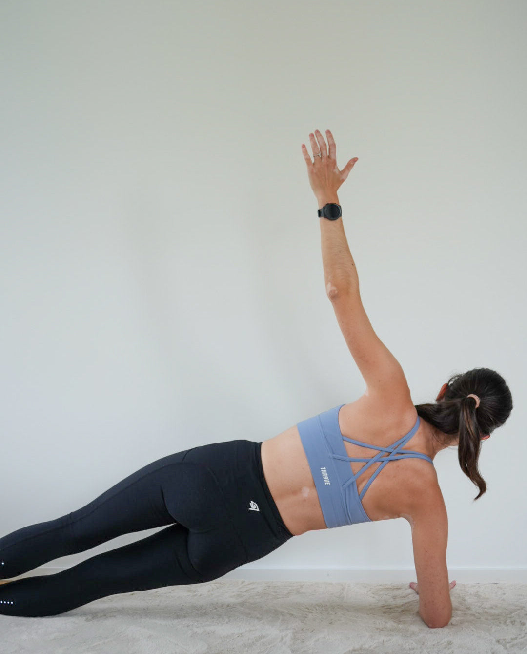 A woman in a light blue sports bra and black leggings performs a side plank stretch on a light-colored floor against a plain wall.