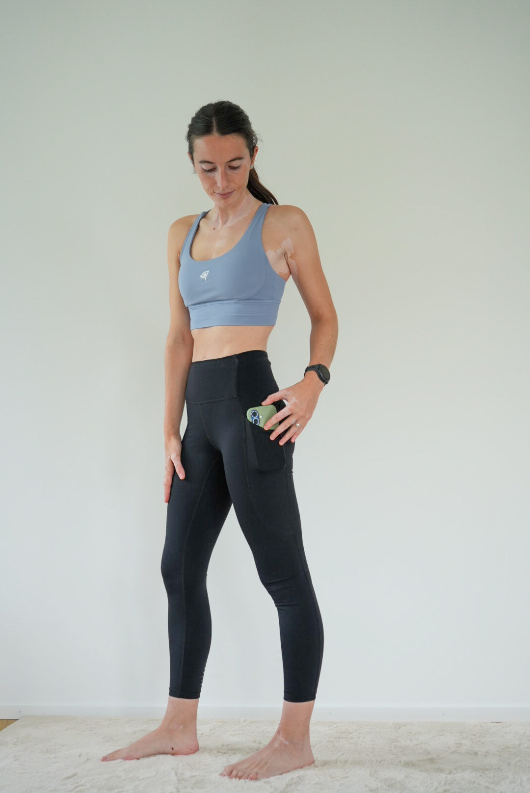 A woman stands barefoot in a studio setting, wearing a light blue sports bra and black high-waisted leggings with a side pocket holding a phone.