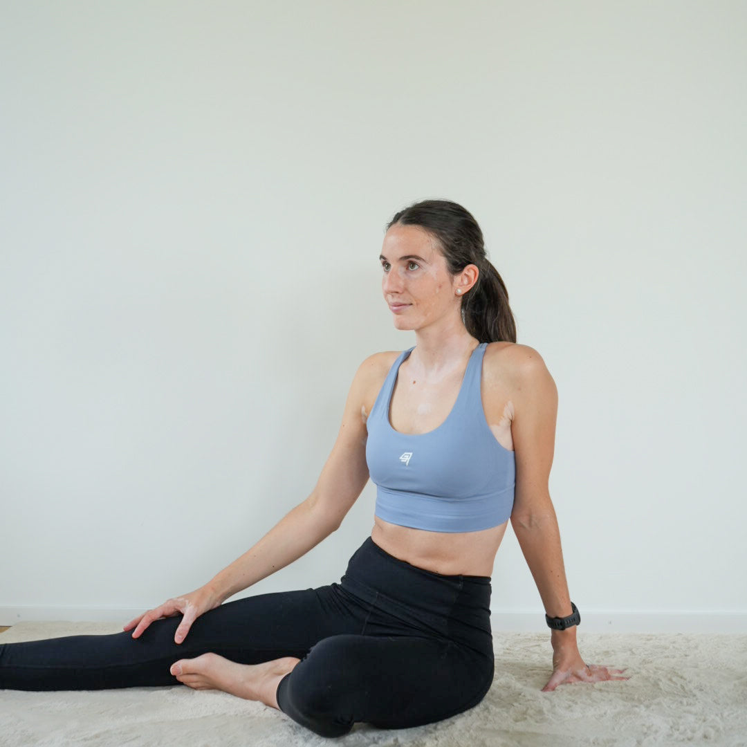A woman sits on a light beige carpet against a plain off-white wall, wearing a light blue sports bra and black leggings.