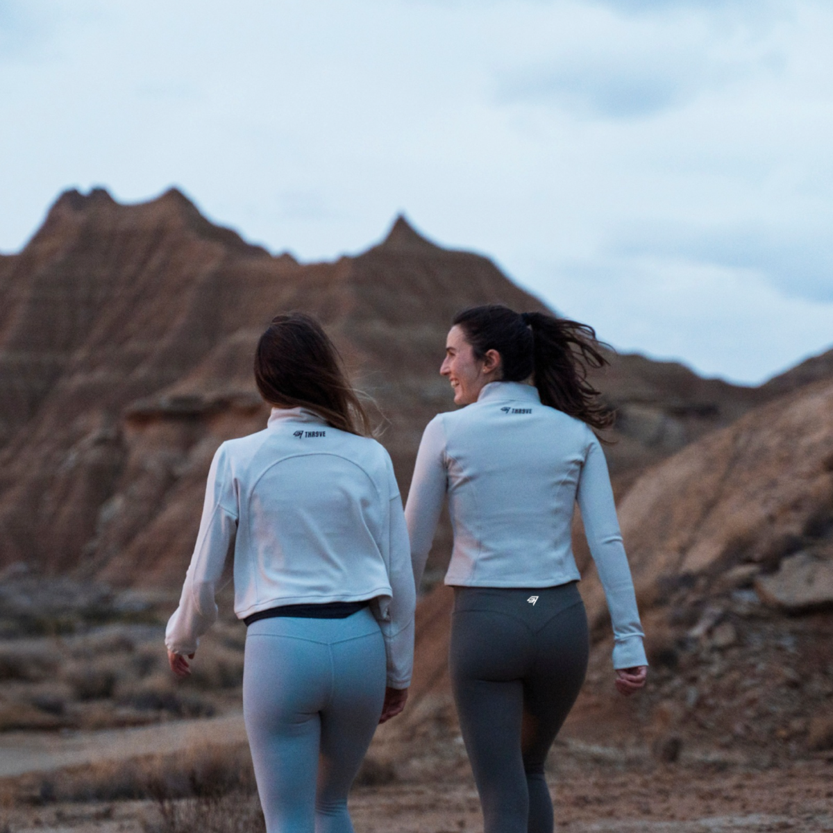 Two women walking hand in hand away from the camera in a desert landscape, wearing matching light gray long-sleeve athletic tops and leggings.