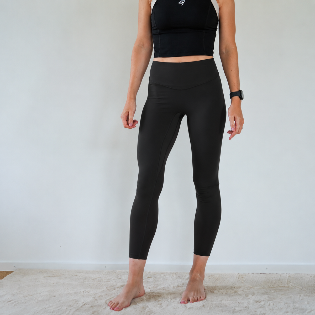 A woman modeling black high-waisted 9/8-length leggings and a black sports bra against a plain white wall and light rug.