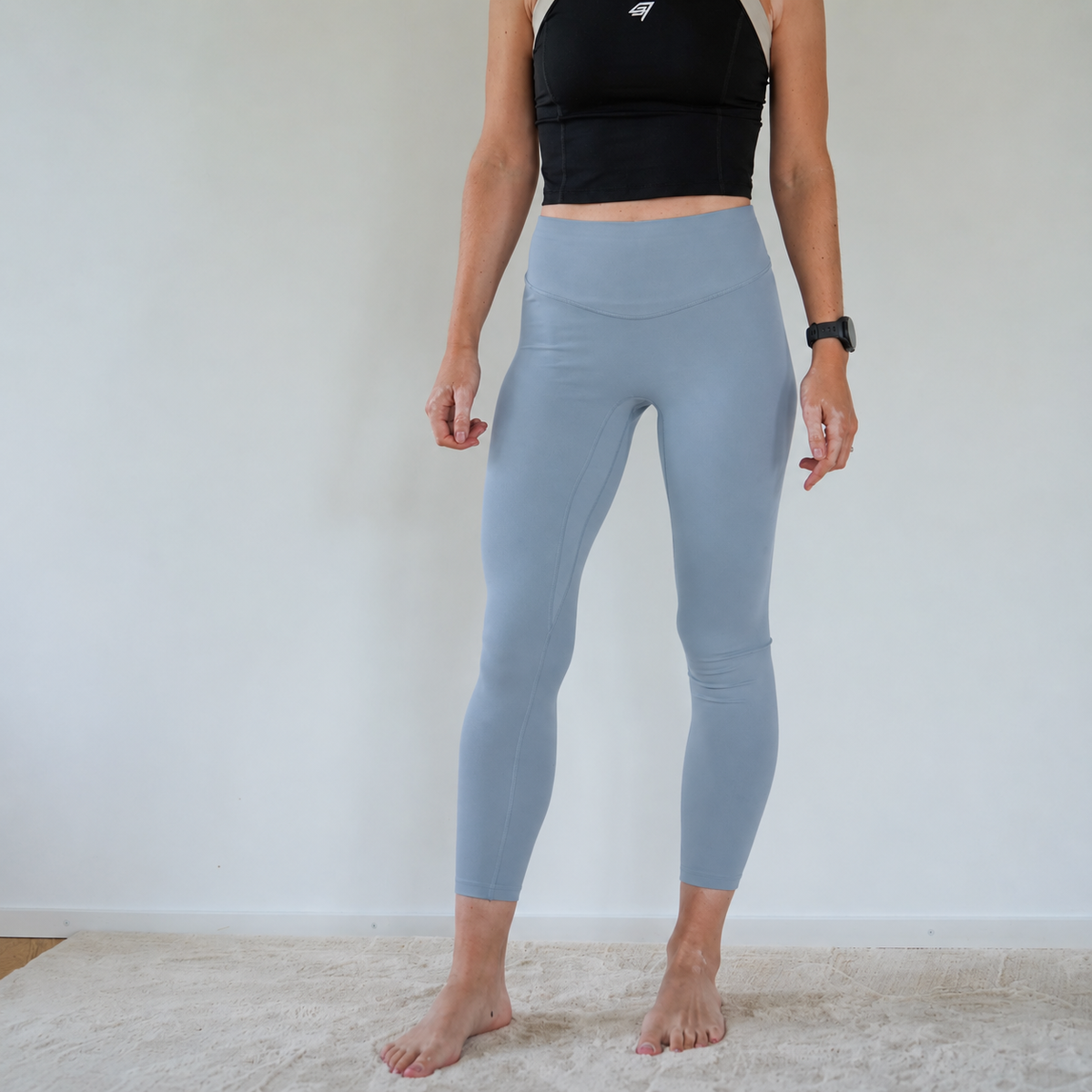 A person modeling high-waisted, ankle-length leggings in a dusty light blue-gray color, paired with a black sports bra, standing barefoot on a light-colored rug against a plain white wall.