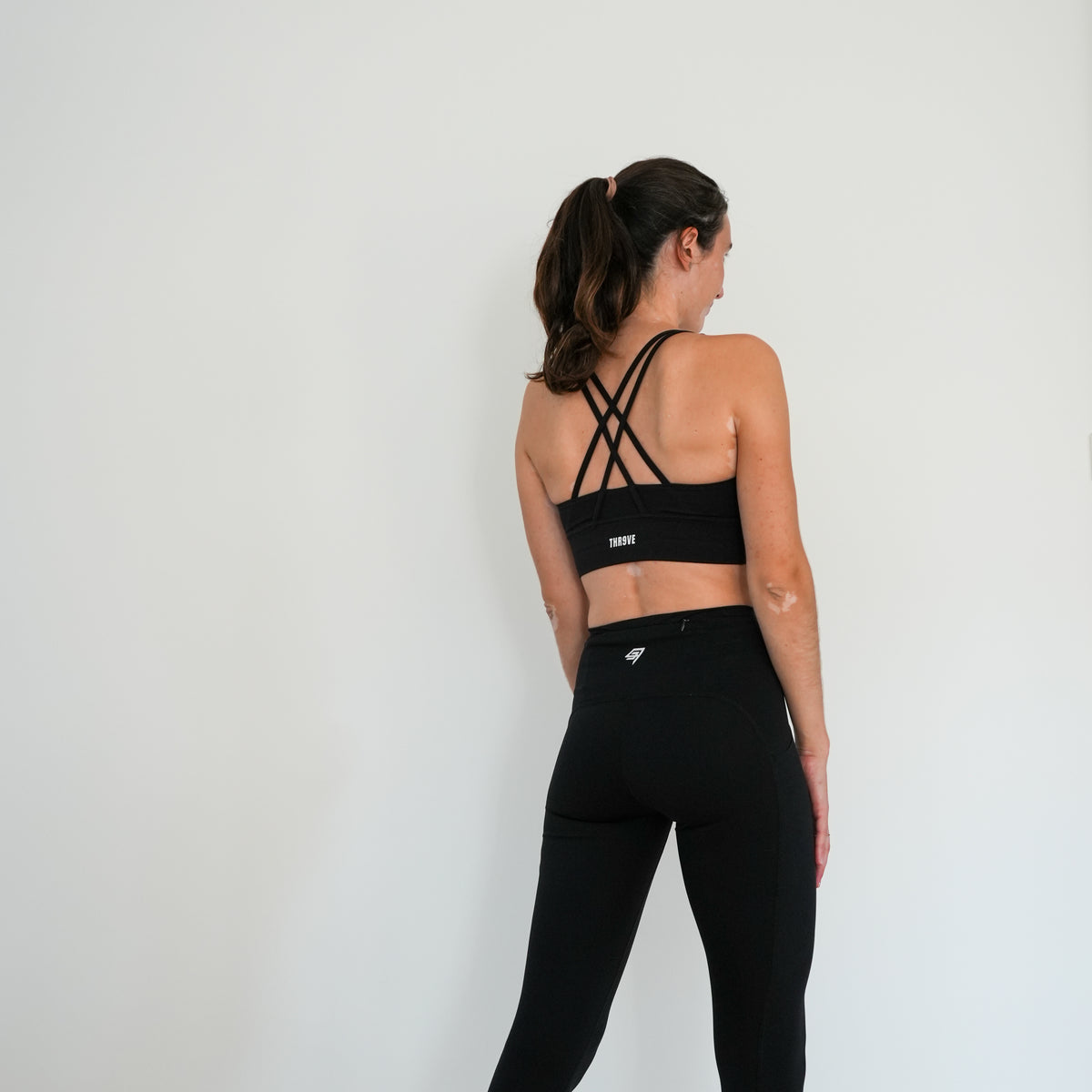 A woman is shown from behind wearing a black sports bra with criss-cross back straps and black leggings, standing against a plain white wall.