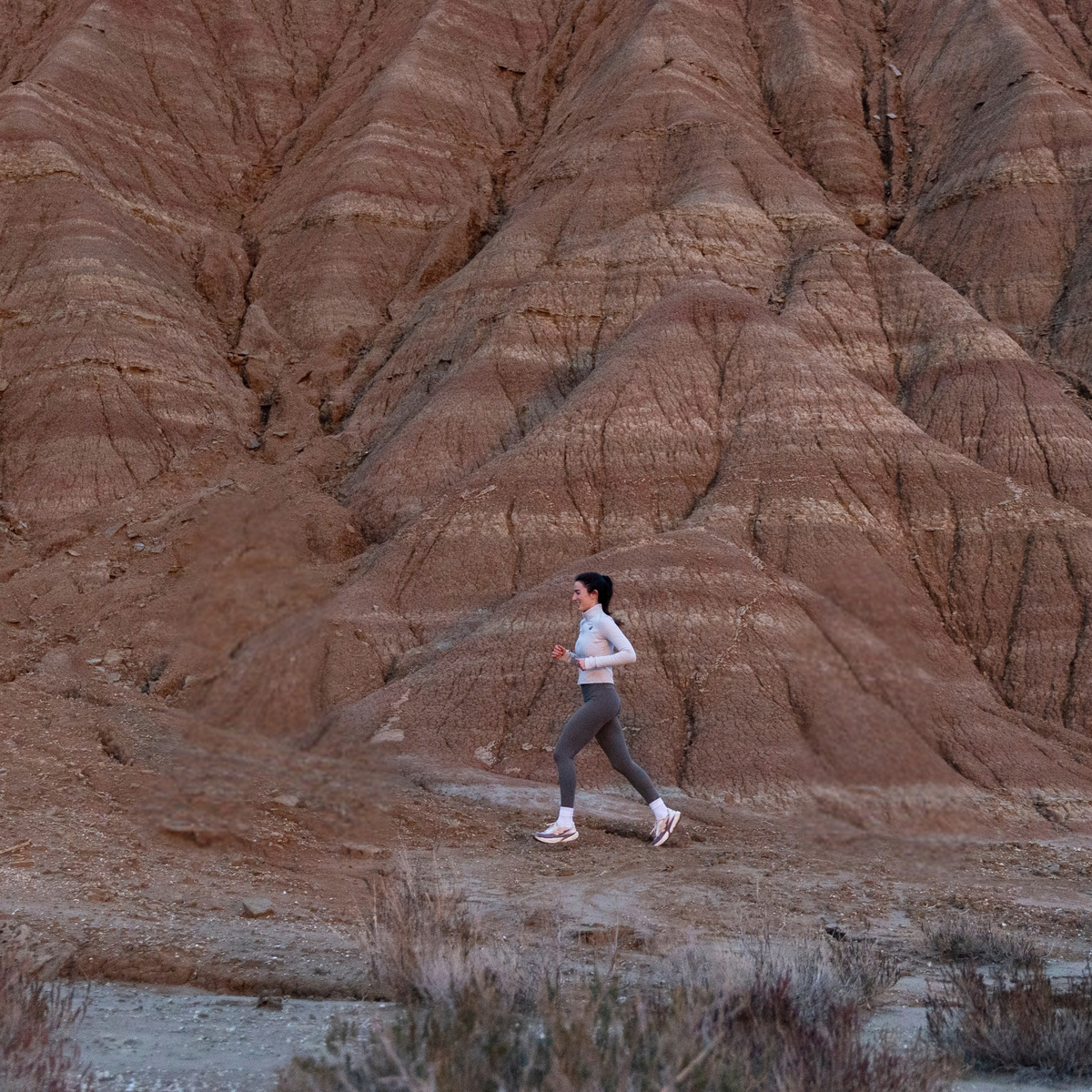 A woman jogging outdoors on a dirt path in front of large reddish-brown badlands rock formations.