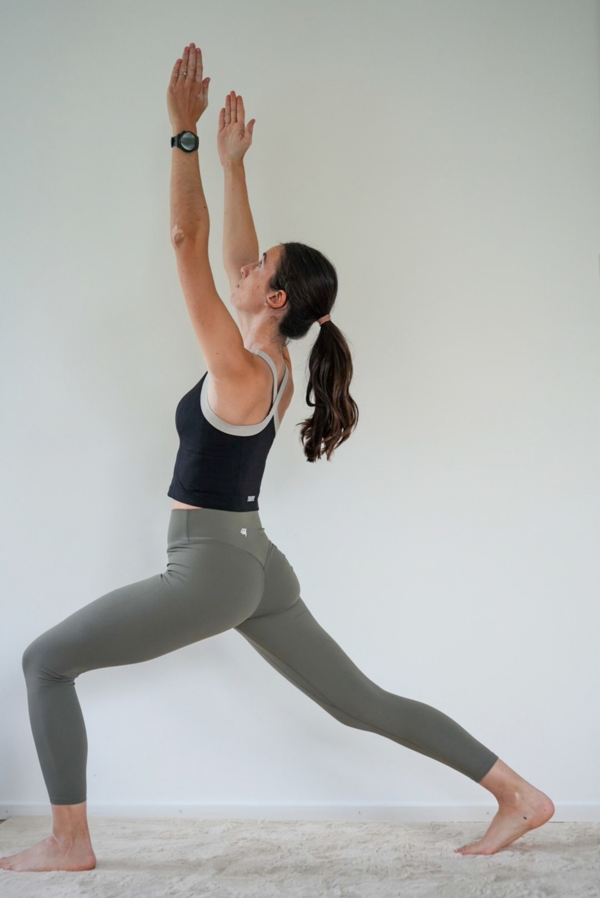 A woman in a yoga pose wearing a black sports bra and olive green leggings, standing against a plain light gray wall.