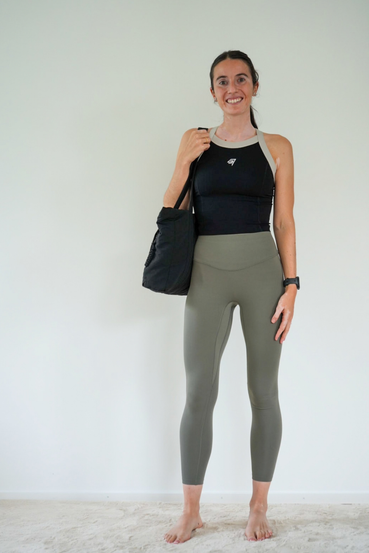 A woman stands barefoot against a plain white wall, wearing a black and gray athletic tank top and olive green high-waisted leggings, while carrying a black tote bag.