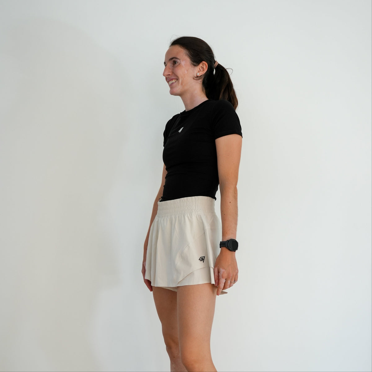 A woman in a black short-sleeve athletic top and a light beige athletic skirt stands in profile against a plain white wall, wearing a black smartwatch.