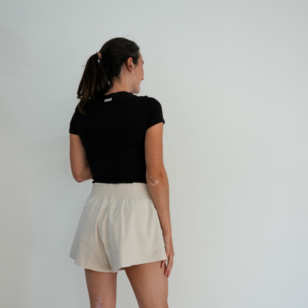 A woman is shown from behind wearing a black short-sleeve t-shirt and cream-colored shorts against a plain white wall.