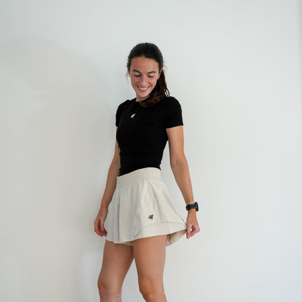 A woman modeling a black short-sleeve athletic t-shirt and a beige tennis skirt with a small black logo, standing against a plain white wall.