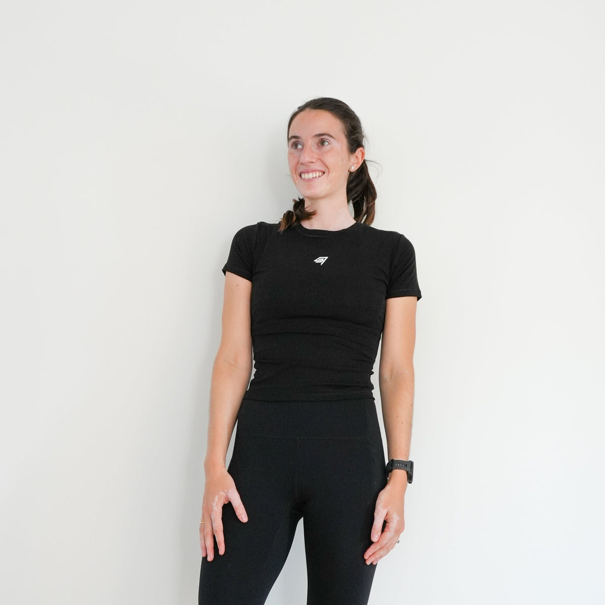 A woman stands barefoot against a plain white wall, wearing a fitted black short-sleeve top and matching black high-waisted leggings.