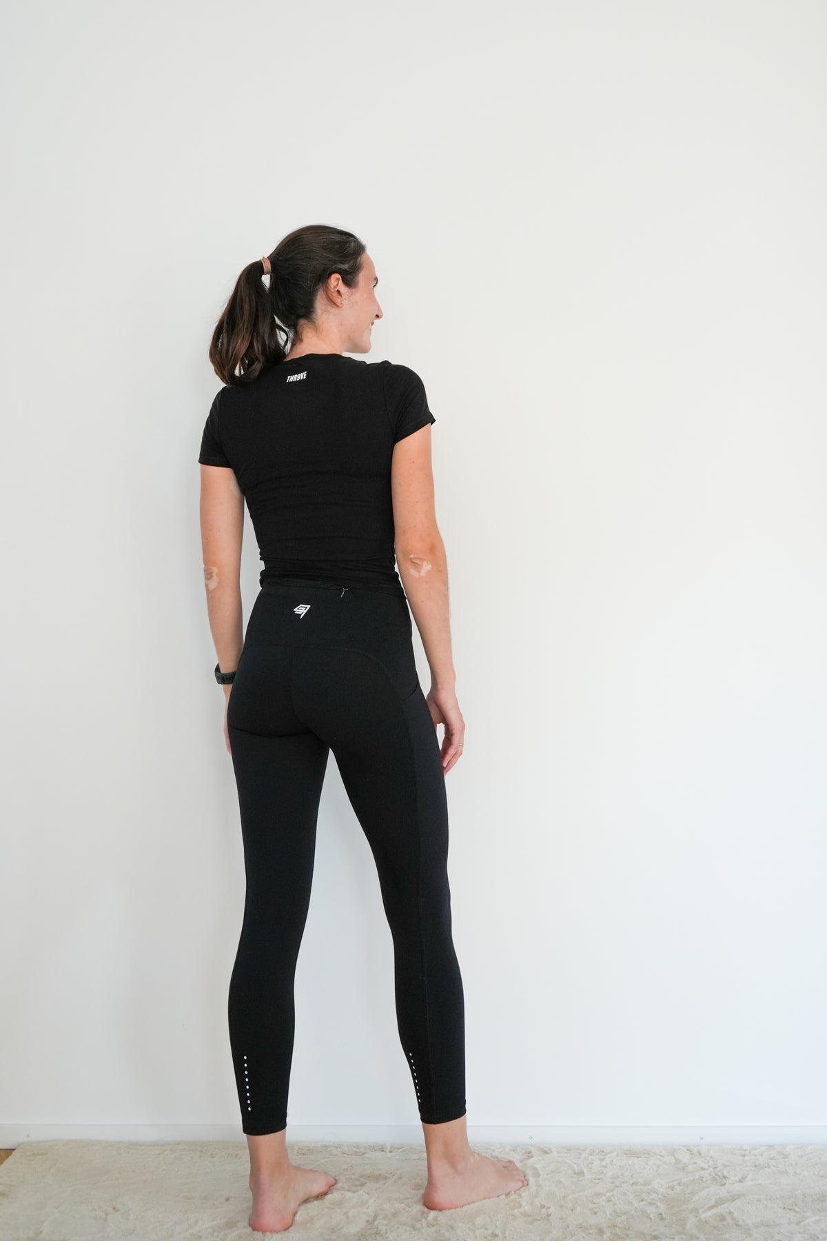 A woman is shown from behind wearing a black short-sleeve t-shirt and black high-waisted leggings with small white geometric accents near the back waistband and ankles, standing against a plain white wall.