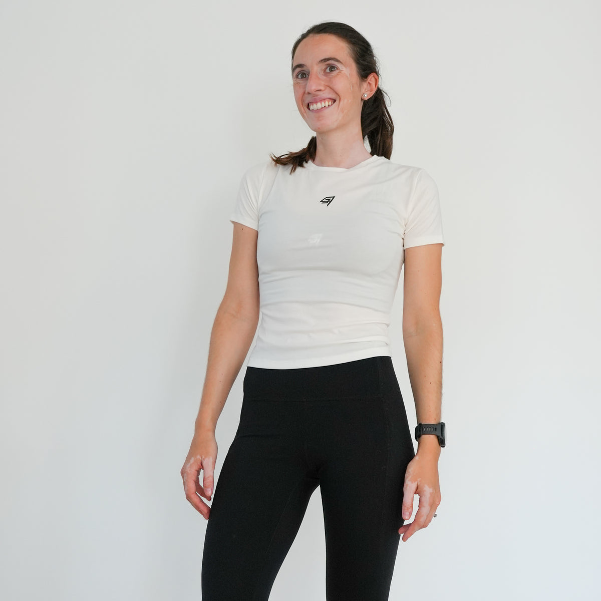 A woman wearing a fitted white short-sleeve athletic t-shirt with a small black logo on the left chest, paired with black leggings, standing against a plain white wall.