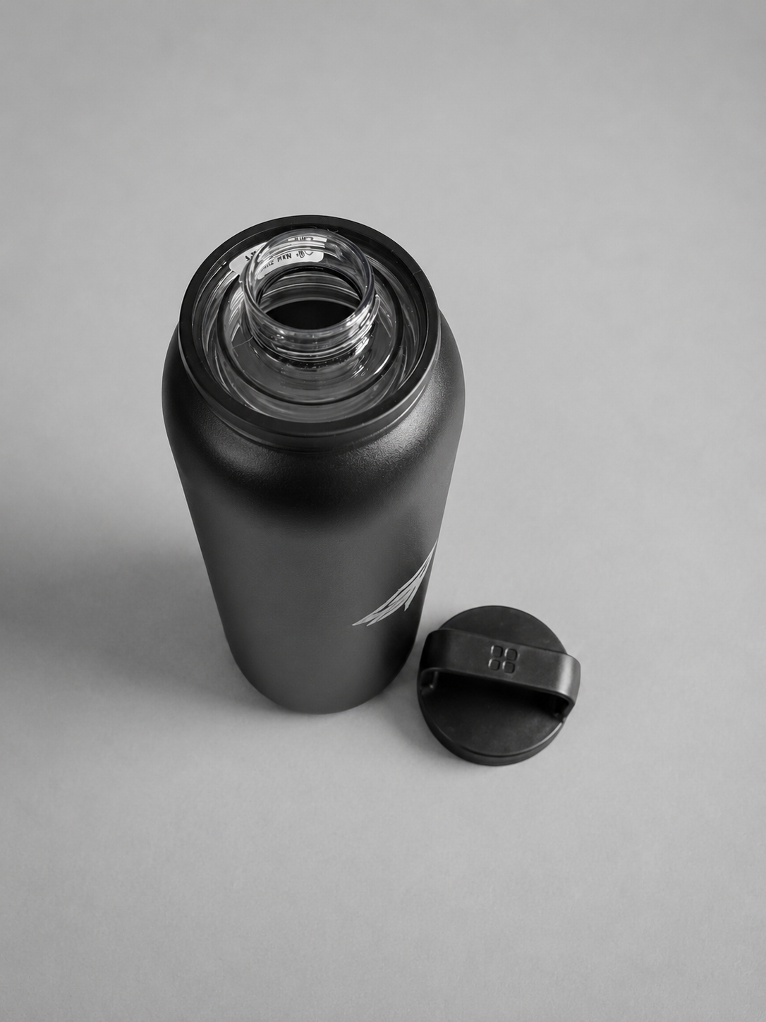 A matte black insulated water bottle with its cap removed and placed beside it on a light gray background.