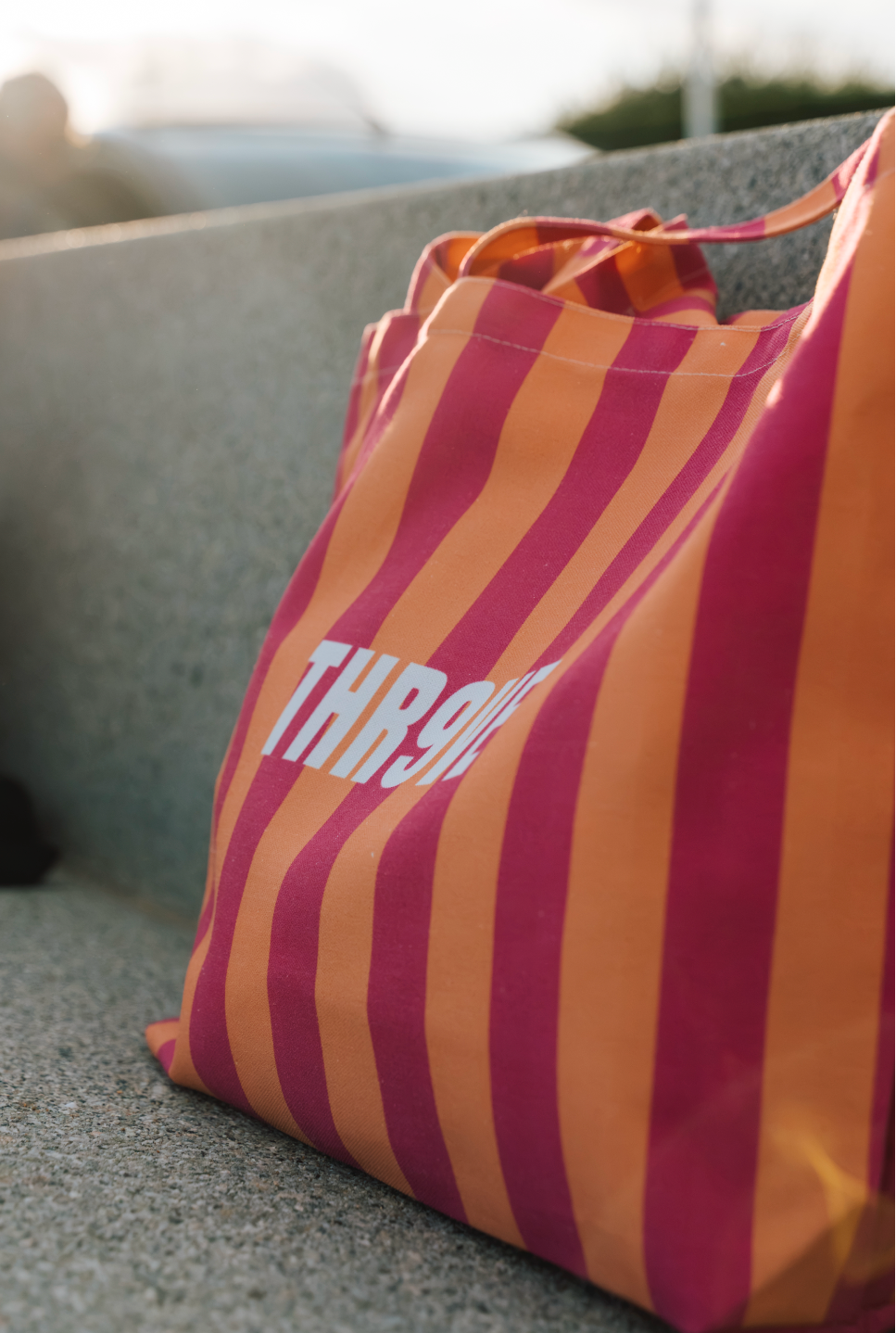 A striped canvas tote bag with alternating orange and magenta pink vertical stripes, featuring the word 'THR9VE' printed in white across the front. The bag is resting on a gray concrete surface with a blurred outdoor background.