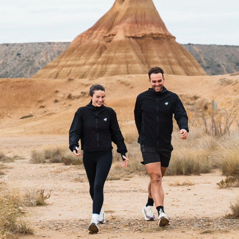 Two people walking in a desert landscape with a unique rock formation in the background.