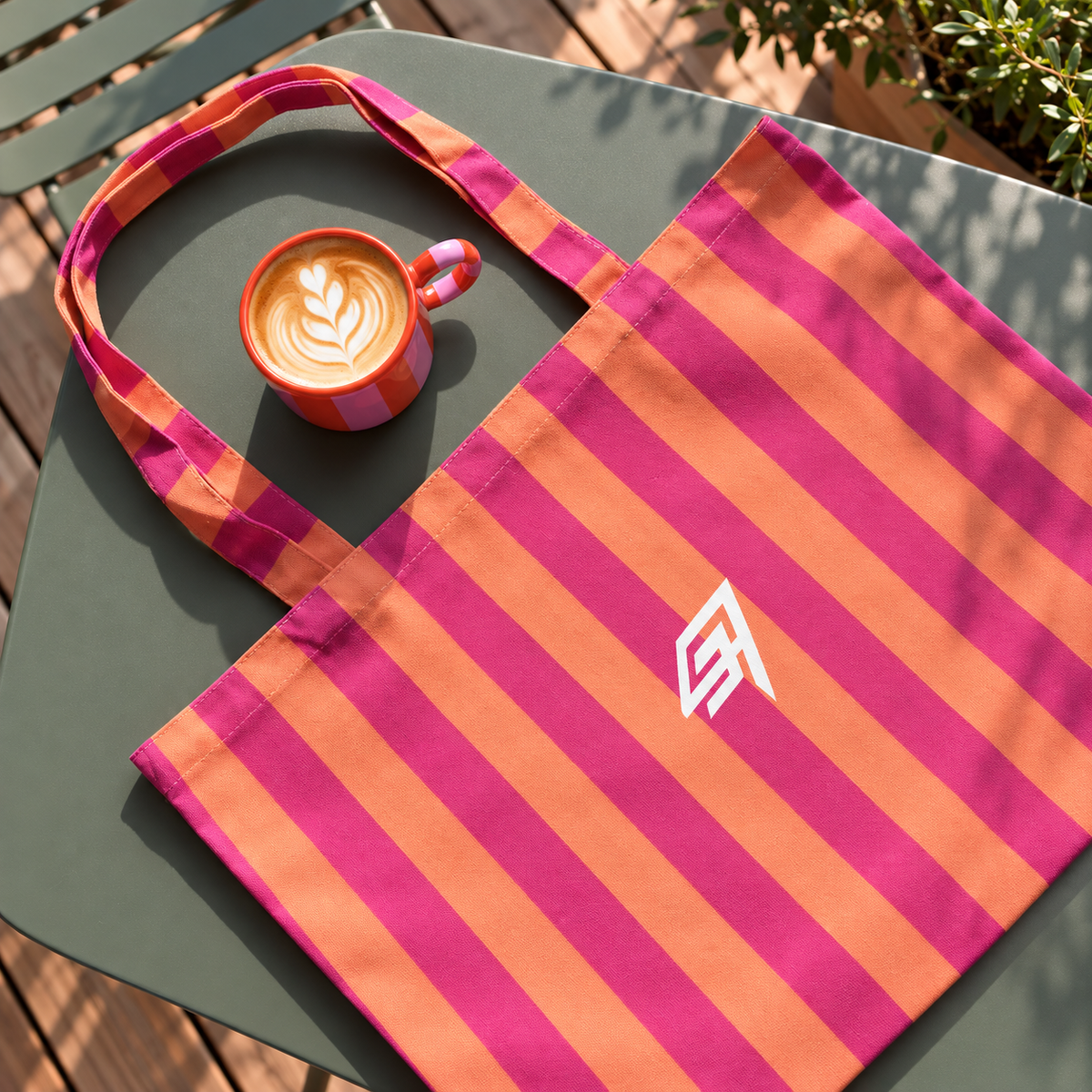 A striped canvas tote bag with bright pink and orange vertical stripes, featuring a white stylized logo on the front. The bag is placed on a dark green outdoor table next to a pink and orange latte cup, with a wooden deck and plants visible in the background.