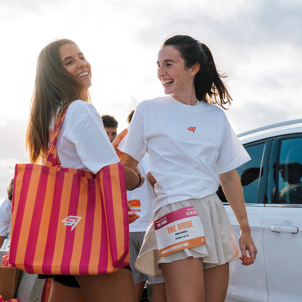 Two young women smiling and laughing outdoors near a white car, wearing matching white athletic T-shirts with a small red logo. One woman carries a large pink and orange striped tote bag.